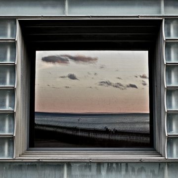 A serene ocean sunset viewed through an industrial-style glass window frame, with pink and gray clouds over the water.