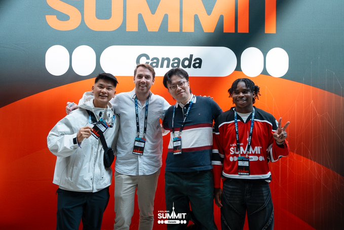 Four men pose together at a 'Canada' 'SUMMIT' event, wearing lanyards and casual attire against a branded orange and gray backdrop.