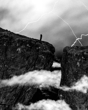 A black and white image showing a tiny figure standing on the edge of a cliff, with lightning streaking across the dark sky in the background.
