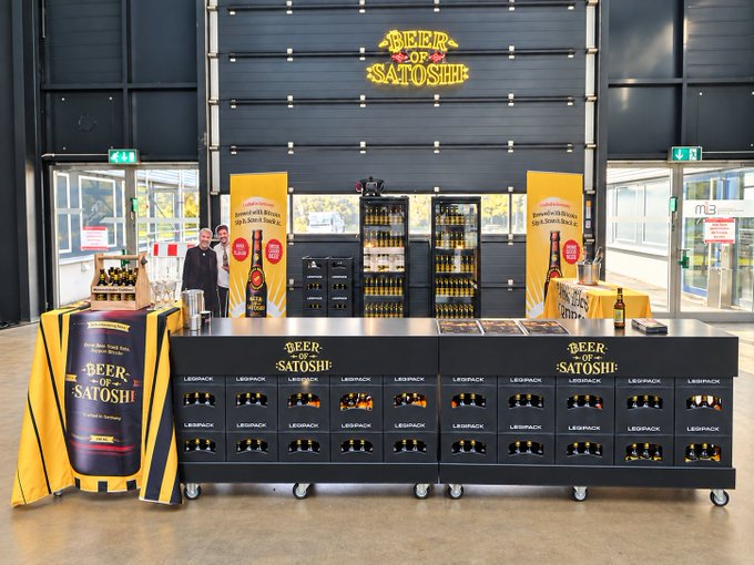 A retail display for 'Beer of Satoshi' featuring branded refrigerators stocked with bottles, a wooden barrel table with a yellow tablecloth, and two people standing in the background.