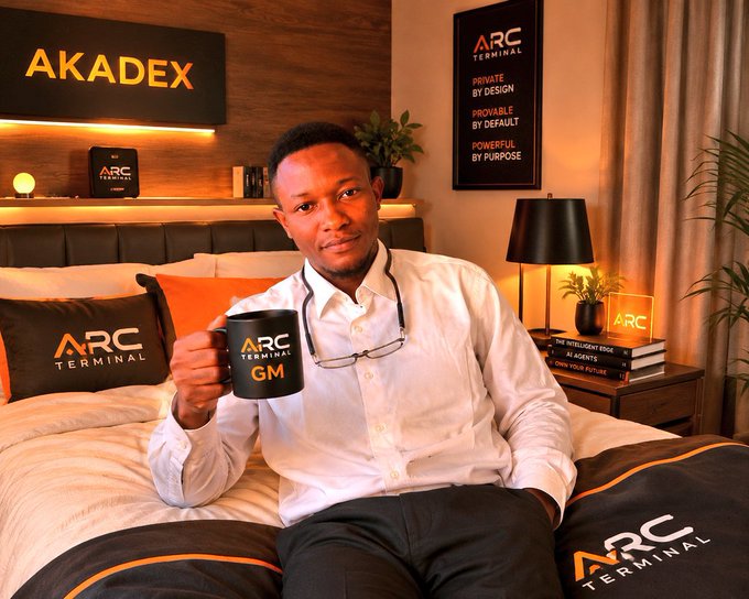 A man sits on a bed in a room with 'AKADEX' and 'ARC TERMINAL' branding, holding a black mug labeled 'ARC TERMINAL GM' with his glasses hanging from his shirt.