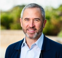 A professional headshot of a bearded, gray-haired man in a dark blazer and light blue shirt, smiling outdoors against a green background. The image appears to represent an industry expert or commentator sharing bullish long-term price predictions for XRP, suggesting the asset could grow exponentially over the next decade.