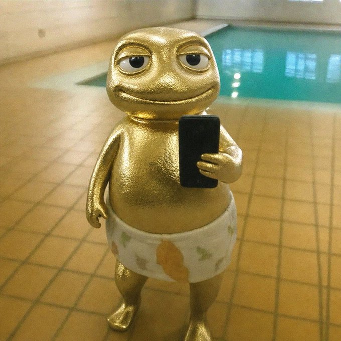 A gold-painted, humanoid statue with bulging eyes and a slight smile stands on a tiled floor near a swimming pool, wearing patterned underwear and holding a black smartphone.