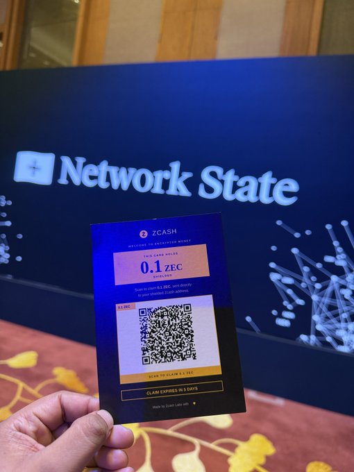A hand holding a Zcash (ZEC) claim card showing a QR code for 0.1 ZEC, with a screen in the background displaying 'Network State' branding and a network graphic.