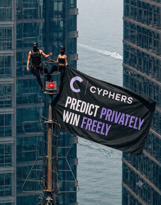 Two figures in dark tactical-style outfits with masks stand atop a metal tower between high-rise buildings, unfurling a large black banner reading 'CYPHERS - PREDICT PRIVATELY, WIN FREELY' over a body of water.