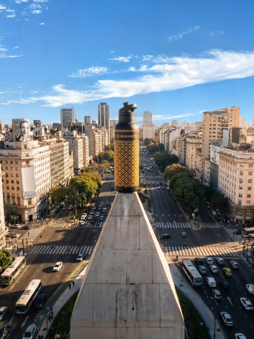 The image shows a cityscape with tall buildings, including one that has a large bottle mounted on top of its roof. The bottle appears to be a perfume bottle or a water dispenser, possibly used for advertising purposes. In addition to the bottle, there are several cars and buses visible in the scene, indicating busy traffic conditions within the city. A bus stop is also present near one of the buildings. The image seems to be related to an advertisement campaign promoting a product or service, as it features a prominent perfume bottle on top of a building amidst the urban environment.