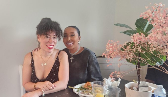Two women pose together at a dining table with a meal and drinks; one has curly hair and wears a polka dot top, while the other has dark hair and wears a black top, with a decorative pink cherry blossom plant beside them.