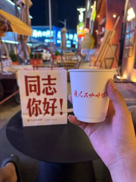 A hand holds a white paper cup in the foreground, while a sign in the background displays the Chinese text "同志你好!" (Hello, comrade!) in red letters. The nighttime street scene, with its illuminated storefronts and colorful signs, complements the friendly greeting featured on the table.
