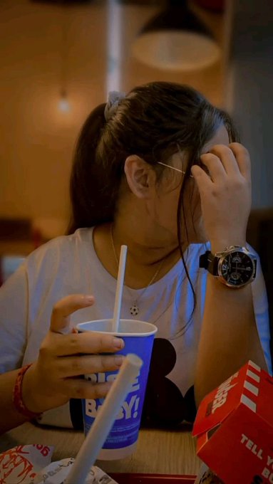 A person with a ponytail and glasses sits at a restaurant table drinking from a blue fast-food cup with a straw, next to a red 'TELL Y...' branded bag.