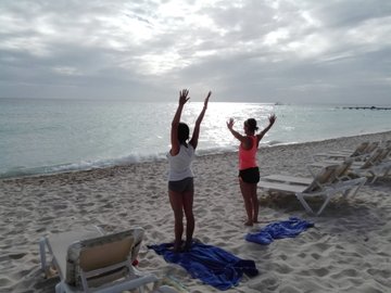 Two people in workout attire stand on a sandy beach with their arms raised toward the ocean under an overcast sky.