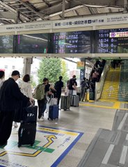 Travelers with luggage stand on a train station platform as digital display boards show departure times.