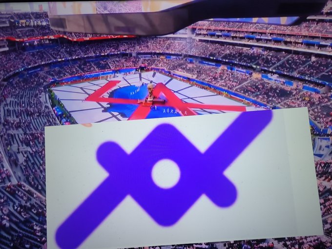 The image shows a large stadium filled with people, possibly at a football game or other sporting event. In the foreground of the photo, there's a white sign that has an unusual purple logo featuring a cross shape and a line through it. The text on the sign reads "Loan protocol in the fifa world cup." This suggests that the image might be related to a discussion about the unique logo or possibly a connection between the event taking place at the stadium and the concept of the loan protocol.