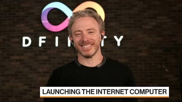 A man with gray hair smiling in front of a DFINITY logo, with a caption reading 'LAUNCHING THE INTERNET COMPUTER'.