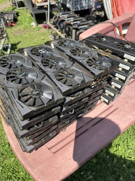 A stack of well-worn graphics cards (GPUs) rests on a wooden table outdoors, their fans and shrouds covered in dust from years of use, with the caption reflecting on the hardware's long mining service since 2017 and its retirement during a spring cleaning.