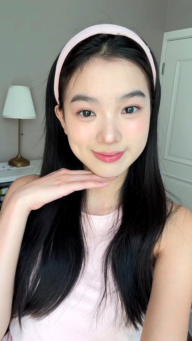 A selfie of a young woman with long dark hair wearing a pink headband and a light pink top, smiling with her hand near her chin.