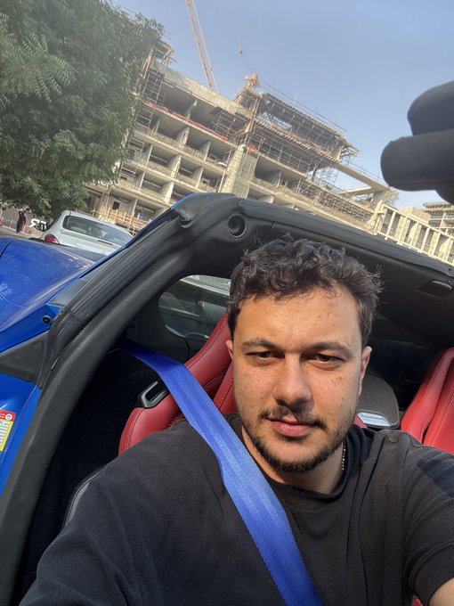 A selfie of a man sitting in the driver's seat of a blue convertible car with a red interior, with a building under construction visible in the background.