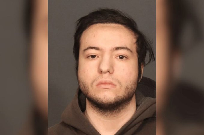 A mugshot-style photo of a young man with dark hair and a brown hoodie against a gray background.