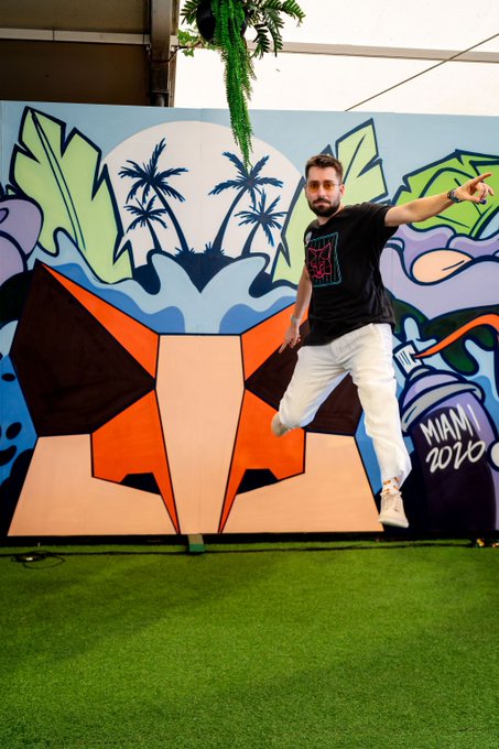 A man jumps enthusiastically in front of a vibrant mural featuring palm trees, a moon, and the text "MIAMI 2020," capturing the energetic spirit of Community Builder Nights being held globally in cities like Dallas, Indonesia, Nigeria, India, and Brazil.