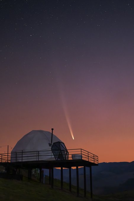 A silhouette of a dome-shaped structure on a wooden platform is set against a vibrant sunset sky featuring a bright comet with a long tail and scattered stars.