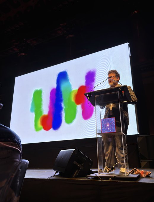 A man wearing glasses standing at a clear acrylic podium on a stage, speaking into a microphone with a large screen behind him displaying colorful abstract artwork in green, blue, red, and purple tones.