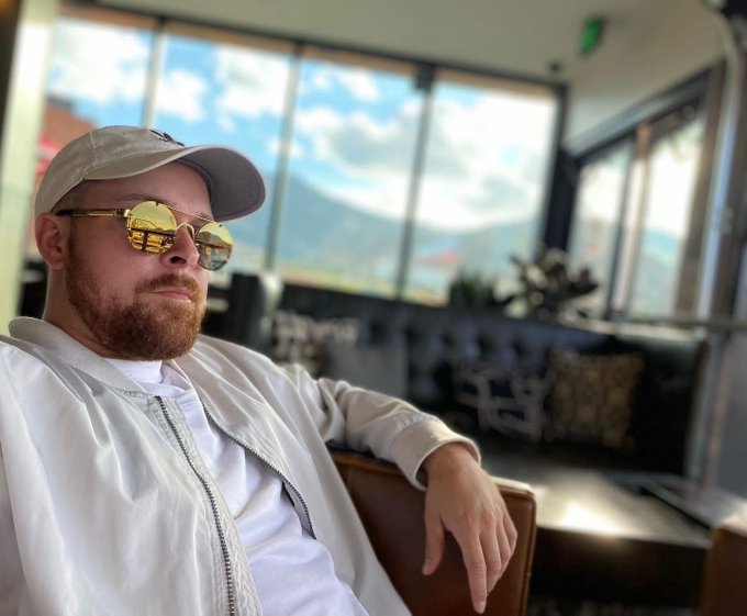 A bearded man wearing a white cap, reflective gold sunglasses, and a cream jacket sits in a chair looking toward the camera.