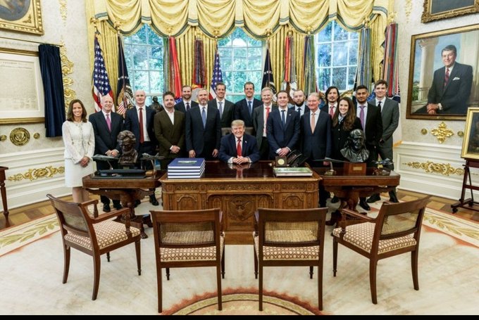 President Donald Trump sits at the Resolute Desk in the Oval Office surrounded by a large group of officials and staff posing for a photograph.