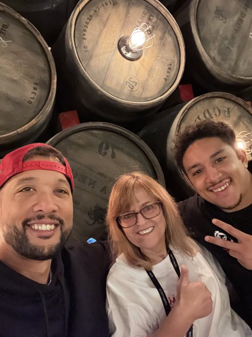 Three people take a selfie in front of stacked wooden barrels, including one labeled "TROUBLE BREWING."
