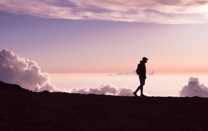 A silhouette of a hiker with a backpack walking along a ridge at sunset, with pastel pink and purple clouds filling the sky above.