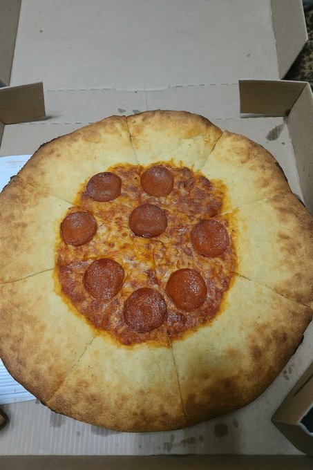 The image features a large pepperoni pizza sitting inside an open cardboard box, which has been placed on top of a table or countertop. The pizza appears to be freshly baked with a golden crust and melted cheese covering the toppings. The text in the image reads "Hey look! A Tezos pizza!". This suggests that the pizza is from a pizzeria known for its unique and creative pizza designs, possibly inspired by the Tezos geometric system used in ancient Greek mathematics.