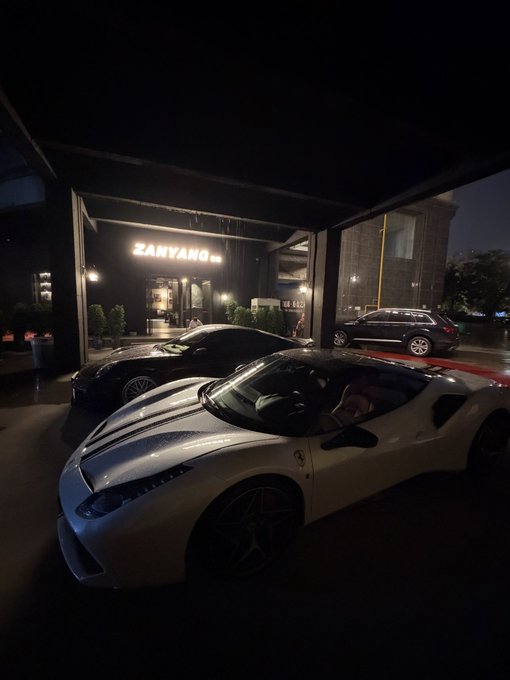 A silver Ferrari with red accents is parked under a covered entrance at night, with the illuminated "ZANYANG" sign visible in the background. The sleek supercar embodies the luxury and exclusivity often associated with successful crypto investments, matching the celebratory tone of the post.