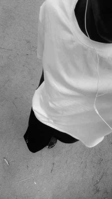 Black and white photograph looking down at a person wearing a flowing white sheer top layered over dark clothing, standing on a concrete surface with small debris scattered around.