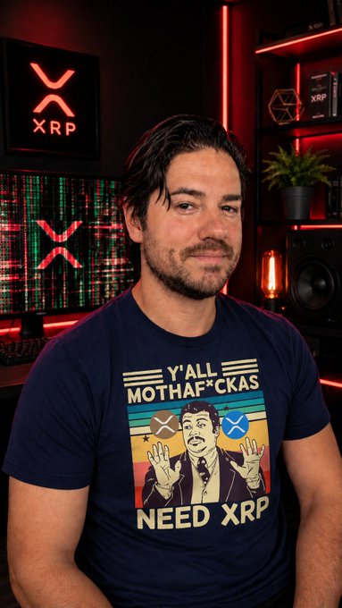 A man with dark hair and a beard wearing a navy 'Y'ALL MOTHAF*CKAS NEED XRP' t-shirt sits in a red-lit room with XRP logos and crypto price charts on the monitors behind him.