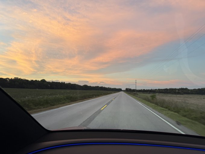 A scenic view from inside a car driving on an empty highway at sunset, featuring a sky painted with vibrant pink and orange clouds over rural fields.