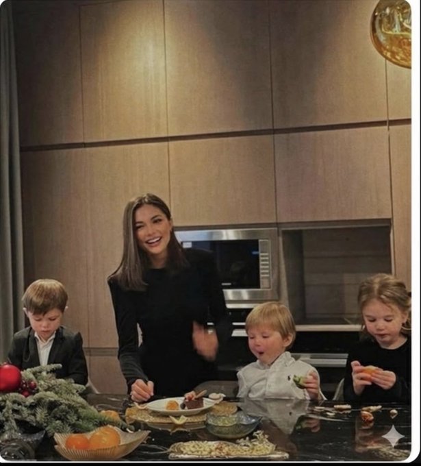 A smiling mother stands behind a kitchen counter with three young children, surrounded by an array of food including oranges, vegetables, and various dishes. The image accompanies a humorous post about family wealth tied to crypto investments, with the woman's cheerful demeanor contrasted against the kids curiously inspecting their snacks, evoking the irony of the joke about explaining crypto gains to children.