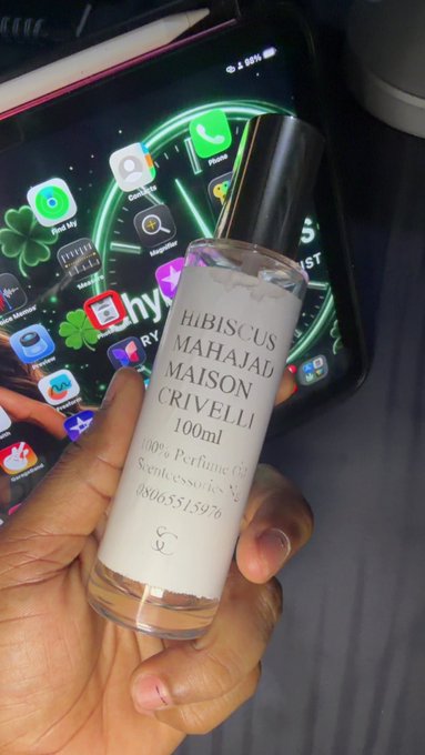 A hand holds a 100ml frosted glass bottle labeled 'HiBiscus Mahajad Maison Crivelli' in front of a smartphone screen displaying app icons.