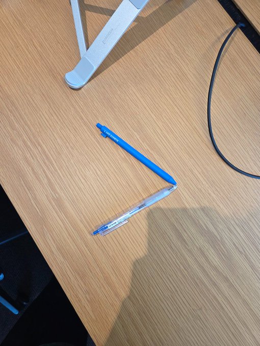 Two blue pens are arranged on a wooden desk with their tips meeting at a chevron angle, alongside a white monitor stand and a black cable. The handwritten caption jokes about crafting a "Bitget Wallet chevron" by placing two pen tips together, playfully suggesting the simple shape resembles the wallet's logo.