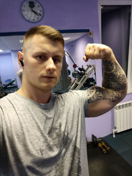 A selfie of a young man with short blonde hair wearing a gray t-shirt, flexing his tattooed right arm in a gym with purple walls and exercise equipment visible in the background.