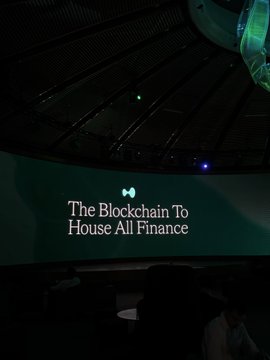 A presentation screen in a dark auditorium displays the text "The Blockchain To House All Finance" in an elegant white font with a small bowtie logo above it.