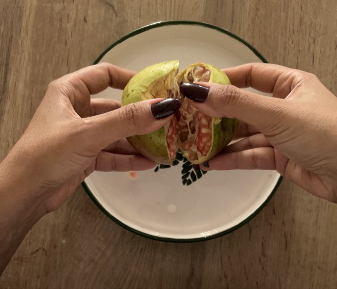 Two hands with dark painted nails are opening a green fruit (appearing to be a walnut or similar) over a white plate with green rim, revealing its interior on a wooden table.