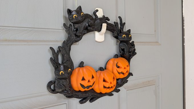 A Halloween-themed decorative wreath hangs on a white door, featuring black cats, twisted branches, and three orange jack-o'-lantern figures.