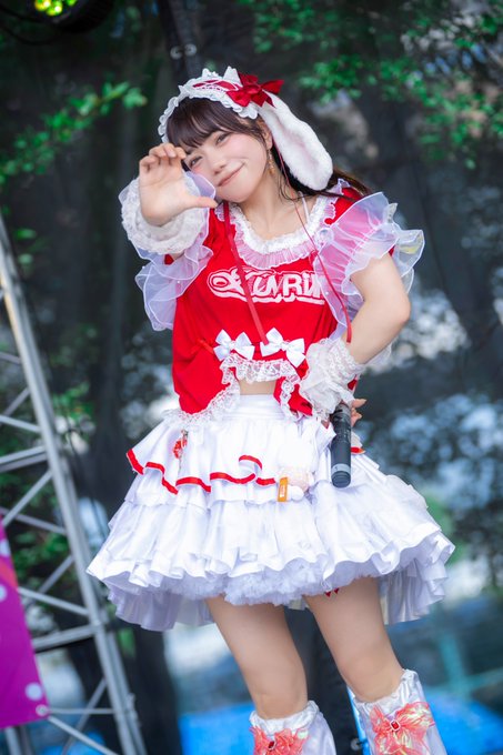 A young woman in a red and white maid-style outfit featuring the text 'CLARE' on her top, posing with a peace sign while holding a microphone at an outdoor event.