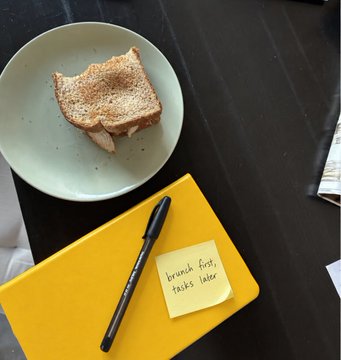 A toasted breakfast sandwich sits on a plate beside a yellow notebook labeled "brunch first, tasks later" with a pen resting on top, humorously illustrating the relatable mood of the "$FLORK being posted again by Binance intern" post—prioritizing leisurely brunch over work responsibilities.