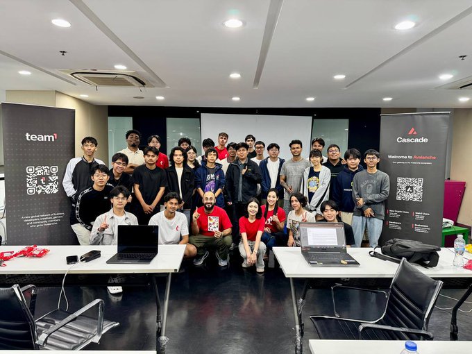 A large group of people pose for a group photo at what appears to be a tech event, with 'team³' and 'Cascade' banners visible, along with laptops on tables in the foreground.
