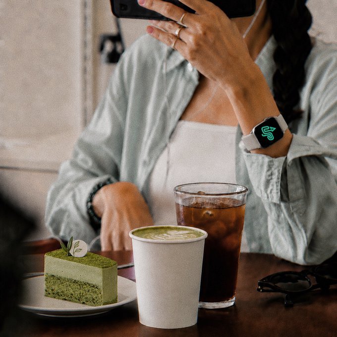 A person at a café with a matcha latte and iced coffee drinks, wearing an Apple Watch showing the time, with a slice of matcha cake on a plate.