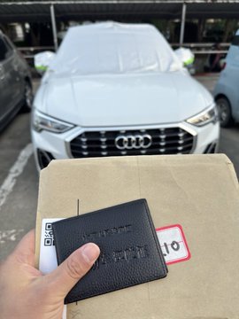 A hand holding a black Audi-branded wallet in front of a cardboard box, with a covered silver Audi SUV parked in the background.