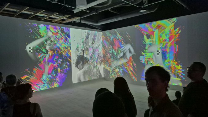 Visitors view large immersive digital art projections featuring colorful glitch-like imagery in a darkened gallery space.