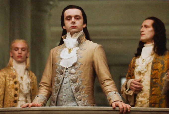 A still from a film showing three men in elaborate 18th-century aristocratic attire, with the central figure in a gold jacket featuring a white ruffled cravat.