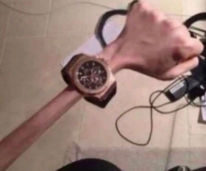 A close-up photo of a person's wrist wearing a wooden watch, holding what appears to be a dumbbell, with their thin arm visible against a tiled floor.