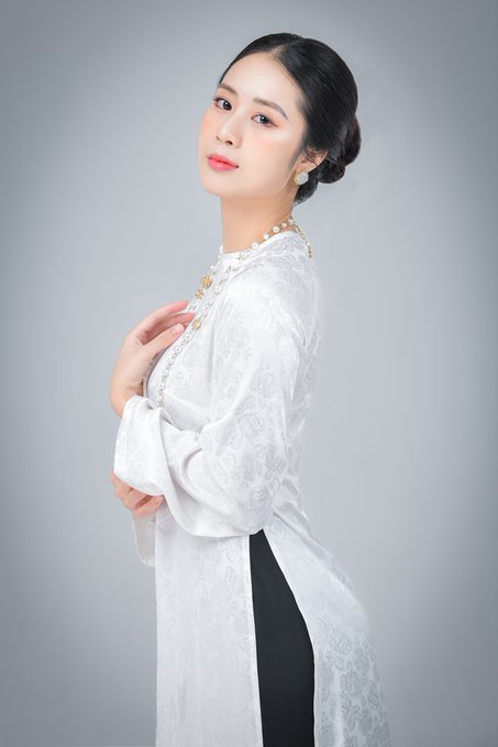 A woman wearing a traditional white Vietnamese áo dài with pearl detailing and a pearl necklace poses gracefully against a grey background.