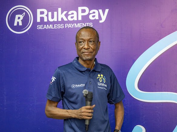 A man wearing a navy blue polo shirt with "RukaPay" and "unicef" logos stands holding a microphone in front of a purple backdrop displaying "RukaPay SEAMLESS PAYMENTS."